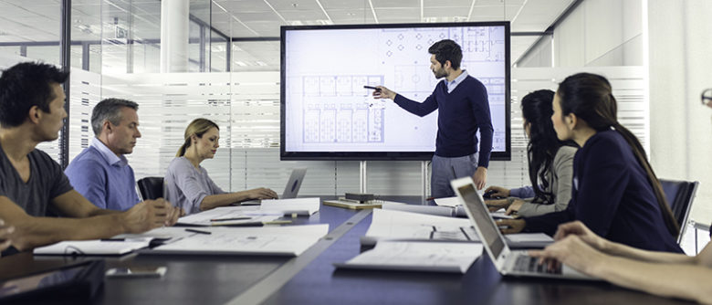 Architect presenting project to a group of managers Engineer standing in front of a presentation screen and pointing to it while explaining details to the audience.