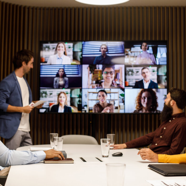 Group of businessmen having a video conference meeting with their colleagues Four businessmen sitting around a conference table in a board room and communicating with their colleagues over a video call. They are discussing business projects and strategies.