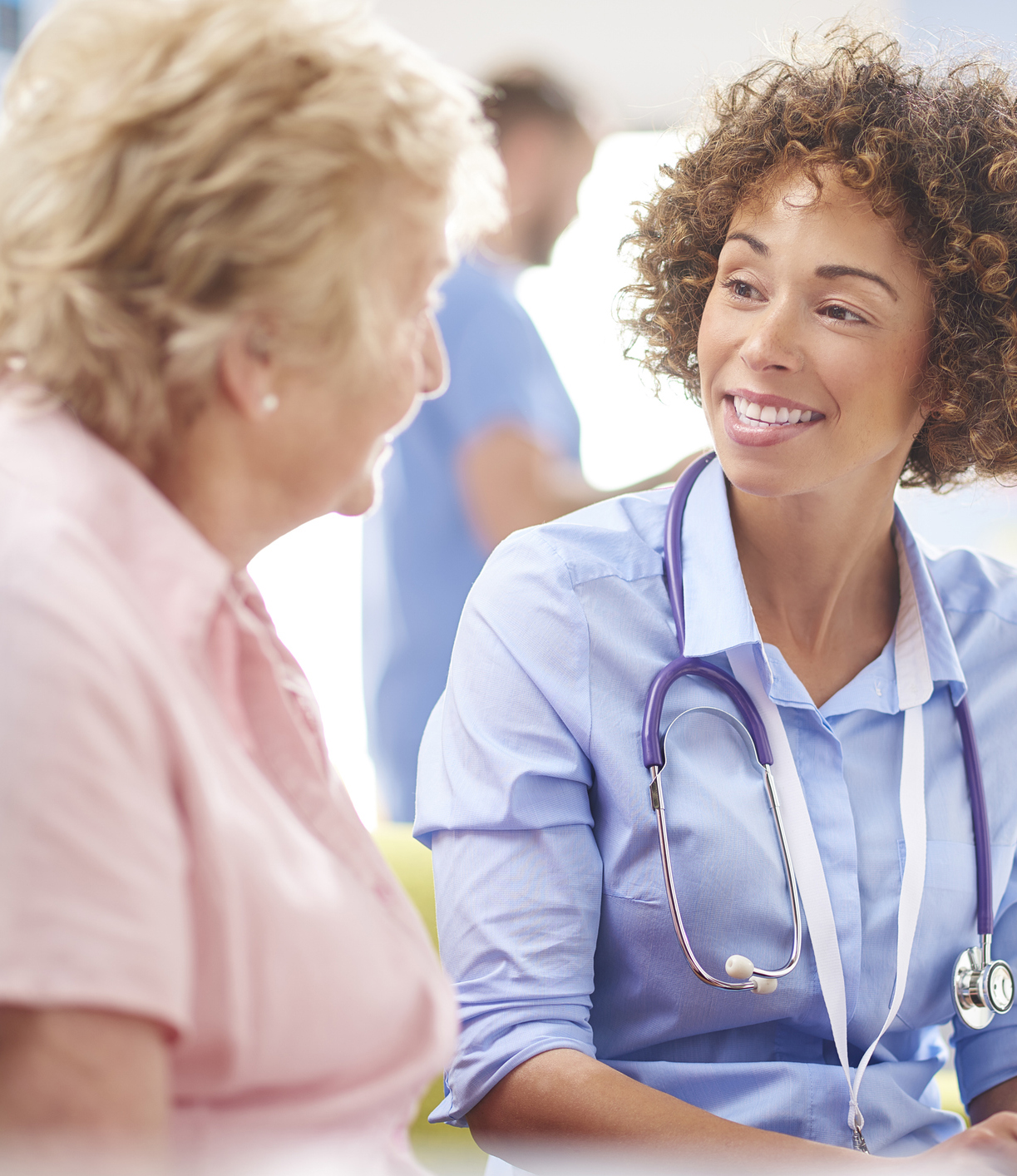 happy doctor with patient a female doctor sits and listens to a senior woman sitting down in a consultation room . They are sitting in a defocussed hospital clinic waiting room . The doctor is wearing no white coat and holding a digital tablet . She is smiling as they chat and a male nurse in scrubs walks past in the background .