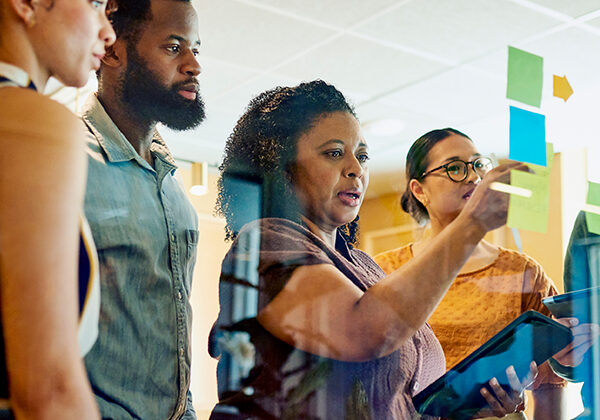 A group of diverse professionals are brainstorming ideas while standing around a glass wall with sticky notes.